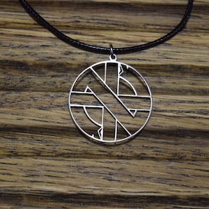 Pode incluir: Um colar com um pingente de prata com um design circular que apresenta um símbolo de anarquia estilizado. O colar está em um cordão preto.