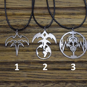 Puede incluir: Tres collares de plata con diferentes colgantes. El primer colgante es un murciélago estilizado con una daga. El segundo colgante es un murciélago estilizado con un globo terráqueo. El tercer colgante es una calavera con un gráfico de latido del corazón.
