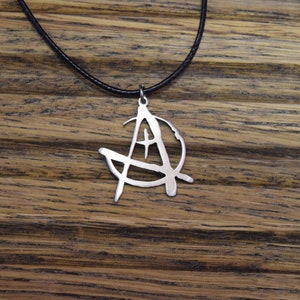 Pode incluir: Um colar de prata com um pingente em forma de símbolo anarquista. O colar está em um cordão preto e está sobre uma superfície de madeira.