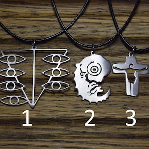 May include: Three silver necklaces with different pendants. The first pendant is a geometric design with multiple eyes. The second pendant is a crescent moon with a face inside. The third pendant is a cross with a figure on it. The necklaces are on a wooden surface.