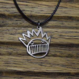 Peut inclure: Un collier en argent avec un pendentif représentant un visage stylisé avec une couronne d'épines. Le pendentif est sur un cordon noir.