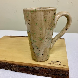 May include: A tall, speckled ceramic mug with a light brown glaze and green speckles. The mug has a rounded handle and a wide base. The mug is sitting on a wooden surface with the text "HAND CRAFTED" and "Molly Mountain Pottery" engraved on the surface.