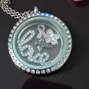 May include: A silver-toned locket necklace with a clear face, featuring the word "Love" spelled out in sparkling crystals. Inside, there's a pink ribbon charm, a cross, and medical symbol. A pink ribbon lies nearby.