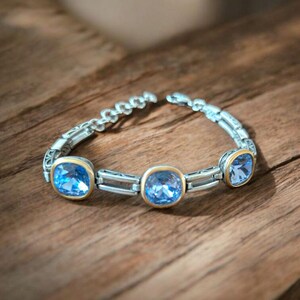 May include: A silver bracelet featuring three large, square, light blue gemstones set in gold-coloured bezels. The bracelet is on a wooden surface, and the links have a decorative pattern.