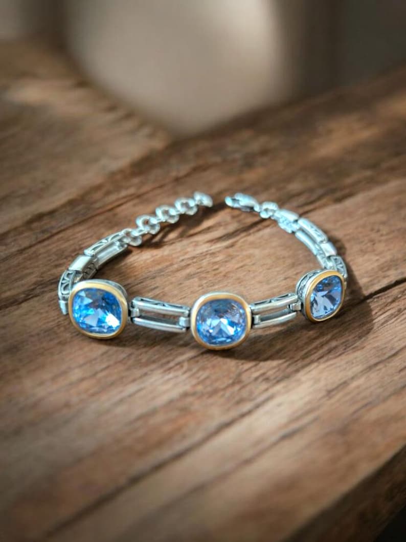 May include: A silver bracelet featuring three large, square, light blue gemstones set in gold-coloured bezels. The bracelet is on a wooden surface, and the links have a decorative pattern.