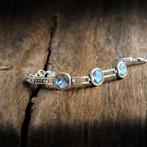 May include: A silver bracelet featuring three square, light blue gemstones set in gold-coloured bezels. The bracelet is displayed on a rustic wooden surface, with a dark background.