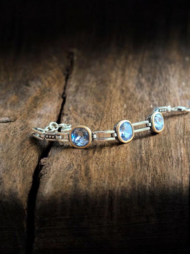 May include: A silver bracelet featuring three square, light blue gemstones set in gold-coloured bezels. The bracelet is displayed on a rustic wooden surface, with a dark background.
