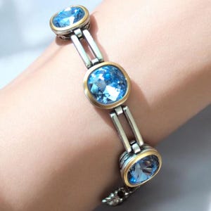 May include: Silver bracelet with three oval, light blue gemstones set in gold-toned bezels. The bracelet has a silver link design and a decorative clasp. The gemstones are faceted, reflecting light.