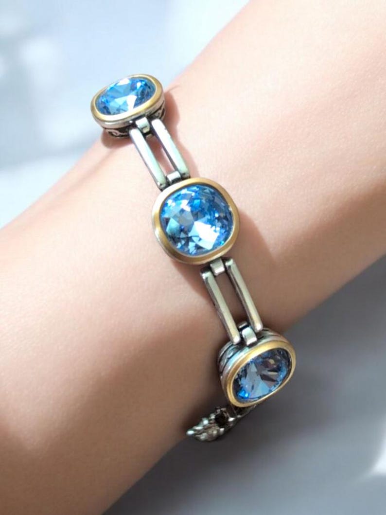 May include: Silver bracelet with three oval, light blue gemstones set in gold-toned bezels. The bracelet has a silver link design and a decorative clasp. The gemstones are faceted, reflecting light.