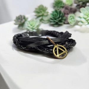 Può includere: Un braccialetto in pelle nera con un design intrecciato e un ciondolo dorato con un simbolo triangolare.