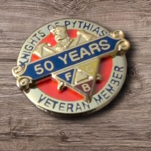 May include: A gold-toned Knights of Pythias Veteran Member pin. The pin features a blue banner with "50 YEARS" and a red and yellow emblem with the letters "F", "C", and "B". The words "Knights of Pythias" and "Veteran Member" are inscribed.