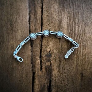 May include: A silver bracelet with square, decorative links and a lobster clasp. The bracelet has a repeating pattern of square centerpieces with a detailed design. The bracelet is displayed on a rustic, wooden surface.