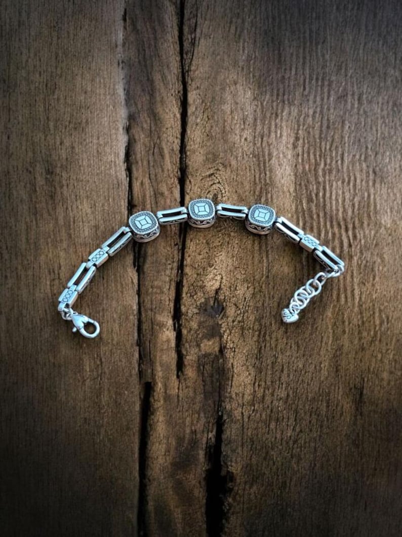 May include: A silver bracelet with square, decorative links and a lobster clasp. The bracelet has a repeating pattern of square centerpieces with a detailed design. The bracelet is displayed on a rustic, wooden surface.