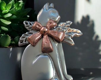 May include: A silver cat figurine with a copper-colored bow and holly leaves. The cat is sitting upright with its head turned to the side.