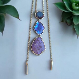 Könnte beinhalten: Eine goldene Kettenkette mit drei violetten und blauen Edelsteinanhängern. Die Anhänger sind facettiert und haben eine geometrische Form.