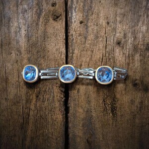 May include: A silver bracelet featuring three large, square, light blue gemstones set in gold bezels. The bracelet is presented on a rustic, dark brown wooden surface, creating a contrast between the jewellery and the background.
