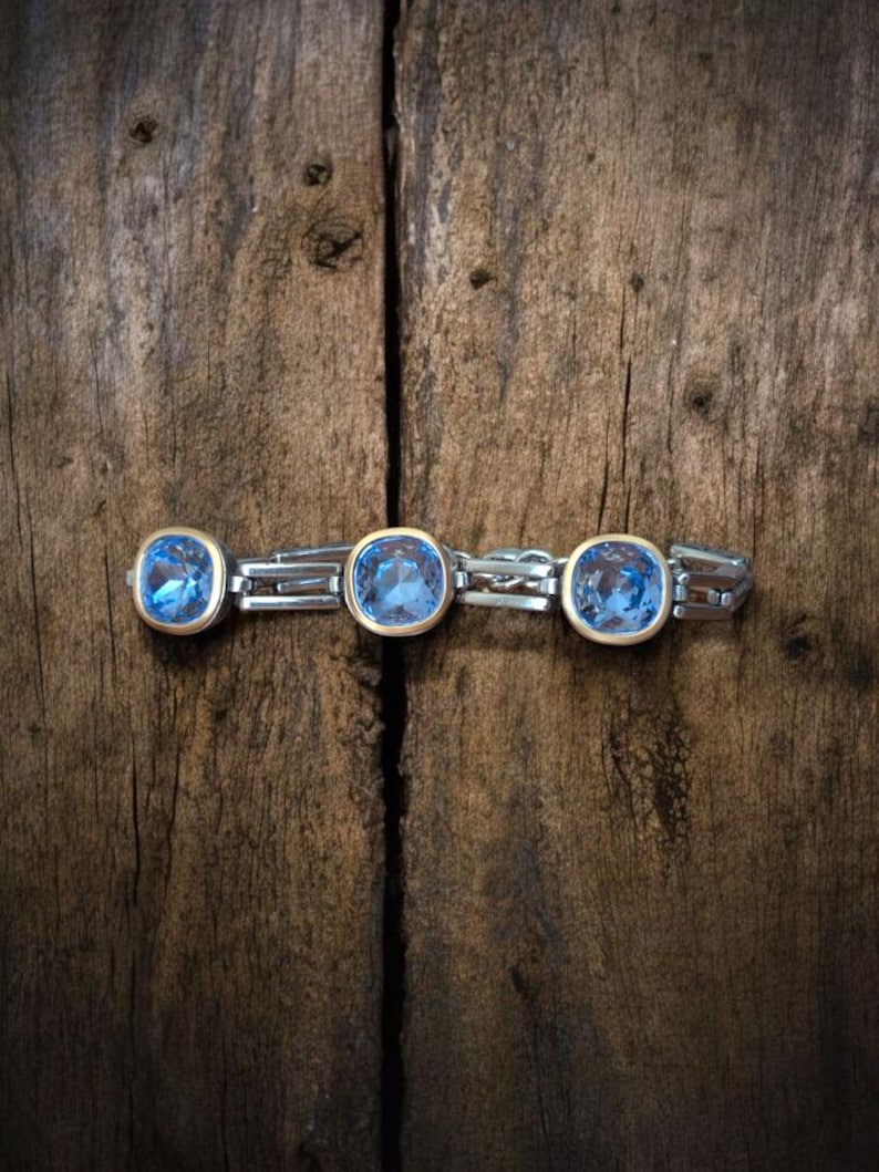 May include: A silver bracelet featuring three large, square, light blue gemstones set in gold bezels. The bracelet is presented on a rustic, dark brown wooden surface, creating a contrast between the jewellery and the background.