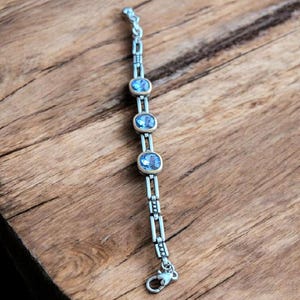 May include: A silver bracelet featuring three oval, pale blue gemstones set in gold. The bracelet has a delicate chain design and a secure clasp. The jewellery is displayed on a wooden surface.