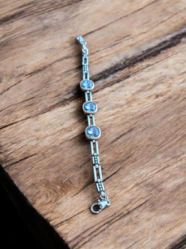 May include: A silver bracelet featuring three oval, pale blue gemstones set in gold. The bracelet has a delicate chain design and a secure clasp. The jewellery is displayed on a wooden surface.