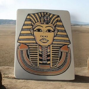 Puede incluir: Una placa decorativa rectangular de color blanquecino con una ilustración estilizada de la cabeza de un faraón egipcio. El tocado del faraón es amarillo y negro, con un detalle de serpiente. La cara es de color canela y el collar es naranja y negro.