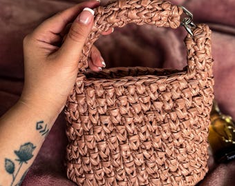 May include: A woven, blush-colored handbag with a rectangular shape and a short handle. The bag features a textured, basket-weave design and a silver-toned metal ring connecting the handle to the bag.