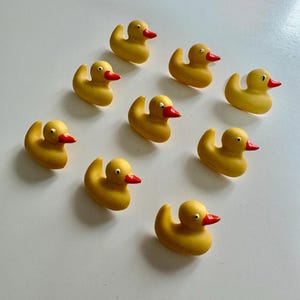 Könnte beinhalten: Zehn gelbe Gummienten mit roten Schnäbeln und schwarzen Augen sind auf einer weißen Oberfläche angeordnet. Diese klassischen Badespielzeuge sind in vielen Haushalten ein vertrauter Anblick.