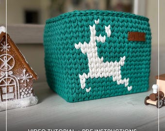 May include: A teal crochet basket with a white reindeer design. The basket is square-shaped and features a small brown leather tag. The text "CROCHET REINDEER BASKET PATTERN" and "VIDEO TUTORIAL + PDF INSTRUCTIONS" are visible.