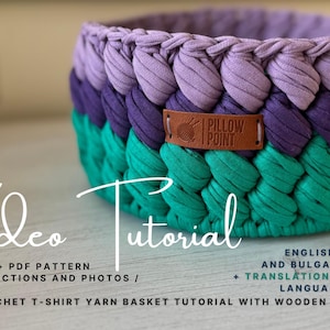 May include: A handmade crochet basket in shades of teal, purple, and lavender. The basket has a braided texture and a leather tag that reads "Pillow Point." The image also includes the words "Video Tutorial" and "Crochet T-Shirt Yarn Basket Tutorial with Wooden Base."