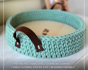 Könnte beinhalten: Ein runder, türkisfarbener Häkeltablett mit hellem Holzsockel und braunem Ledergriff. Das Tablett hat ein strukturiertes Stichmuster und ist für Anfänger konzipiert. Der Text unten lautet "CROCHET TRAY TUTORIAL".