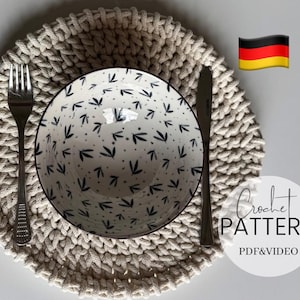 May include: A beige crocheted placemat with a bowl and silverware. The bowl has a white background with a black leaf pattern. A fork and knife are placed on either side of the bowl. The image also includes the text "Crochet Pattern PDF & Video".