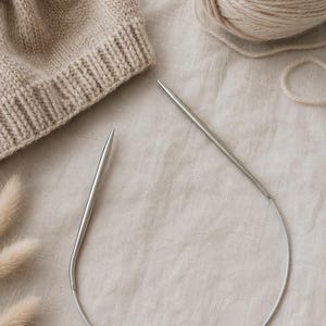 Puede incluir: Un gorro de punto beige, una madeja de lana y un par de agujas de punto circulares plateadas descansan sobre una tela de tono neutro. Las agujas son curvas y están conectadas por un cable flexible, ideal para tejer en redondo.