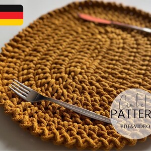 May include: Mustard yellow crocheted placemat with a textured design. A silver fork and knife are placed on the placemat. The words "Crochet Pattern PDF & Video" are in a white circle. A German flag is in the upper left corner.