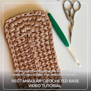 Op de afbeelding: Een rechthoekige gehaakte basis in lichtbruine kleur, met een getextureerd oppervlak. Een groene en witte haaknaald en decoratieve schaar liggen naast de basis. De tekst op de afbeelding luidt "RECTANGULAR CROCHETED BASE VIDEO TUTORIAL".