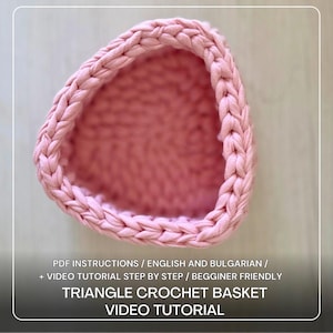 May include: A pink, triangular crochet basket with a braided edge. The basket is made of thick yarn. The image includes text that reads "TRIANGLE CROCHET BASKET VIDEO TUTORIAL".
