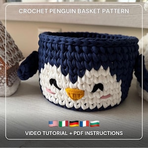May include: A navy blue, white, and yellow crochet penguin basket. The basket has a round shape with a penguin face design on the front. The text "CROCHET PENGUIN BASKET PATTERN" is at the top, and "VIDEO TUTORIAL + PDF INSTRUCTIONS" is at the bottom.