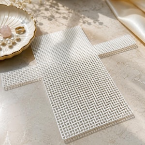 Puede incluir: Una lámina de lona de plástico blanco con un patrón de cuadrícula, ideal para manualidades. La lámina tiene forma de cruz. Un plato decorativo con borde dorado, un anillo y perlas están en el fondo.