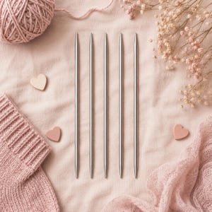 Peut inclure: A set of silver knitting needles on a pink fabric, accompanied by a ball of yarn, a knitted piece, and heart-shaped decorations. The needles are long and thin, perfect for various knitting projects. The composition is soft and delicate.