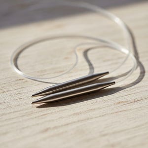 Può includere: Primo piano di un paio di ferri da maglia argentati collegati da un cavo trasparente. Gli aghi sono appuntiti e giacciono su una superficie di legno chiaro. Il cavo forma un anello.