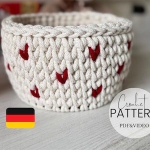 May include: A cream-colored, crocheted basket with red heart accents. The basket has a braided top edge and a round shape. The image includes the text "Crochet Pattern PDF & Video" in a circular design and a German flag.