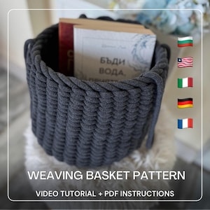 May include: A dark gray woven basket with rope handles, filled with books. The basket is cylindrical and sits on a fluffy white surface. Text at the bottom reads "WEAVING BASKET PATTERN VIDEO TUTORIAL + PDF INSTRUCTIONS."