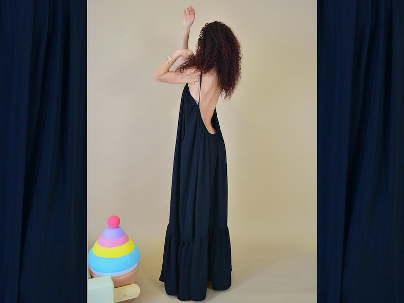May include: A long, flowing black dress with a low back and spaghetti straps. The dress has a tiered, ruffled hemline. The model is wearing the dress with their arms raised. A colorful toy is in the background.