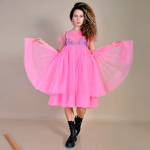 May include: A woman wearing a pink tulle dress with a layered skirt. She is standing on a light brown surface with a wooden baseball bat lying on the floor.