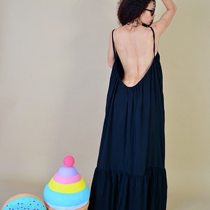 May include: A long, navy blue maxi dress with thin straps and a low back. The dress has a ruffled hemline. Decorative, oversized plush toys of a cupcake, donut, and popsicle are in the foreground.