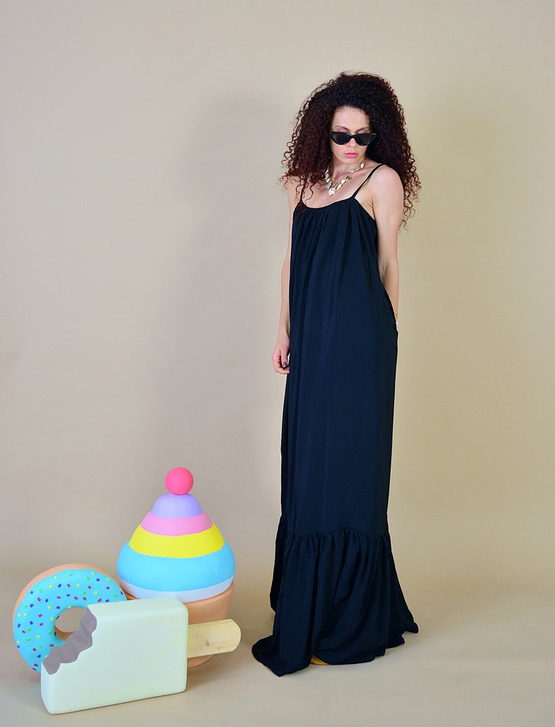 May include: A woman wearing a black maxi dress with spaghetti straps and a ruffled hem. She is wearing sunglasses and a necklace. Decorative oversized ice cream, donut, and popsicle props are in the foreground.
