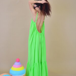 May include: A woman wearing a bright green maxi dress with a low back and ruched detailing. The dress has a tiered skirt and is made of a lightweight fabric. The woman is standing in front of a light brown background.