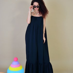 May include: A woman wearing a black maxi dress with spaghetti straps and a ruffled hem. She is wearing sunglasses and a necklace. The dress is long and flowing, and the overall style is casual and chic.