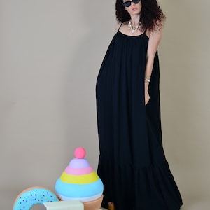 May include: A woman wearing a black maxi dress with spaghetti straps and sunglasses. The dress has a tiered ruffle at the hem. Decorative oversized ice cream, donut, and popsicle props are in the foreground.