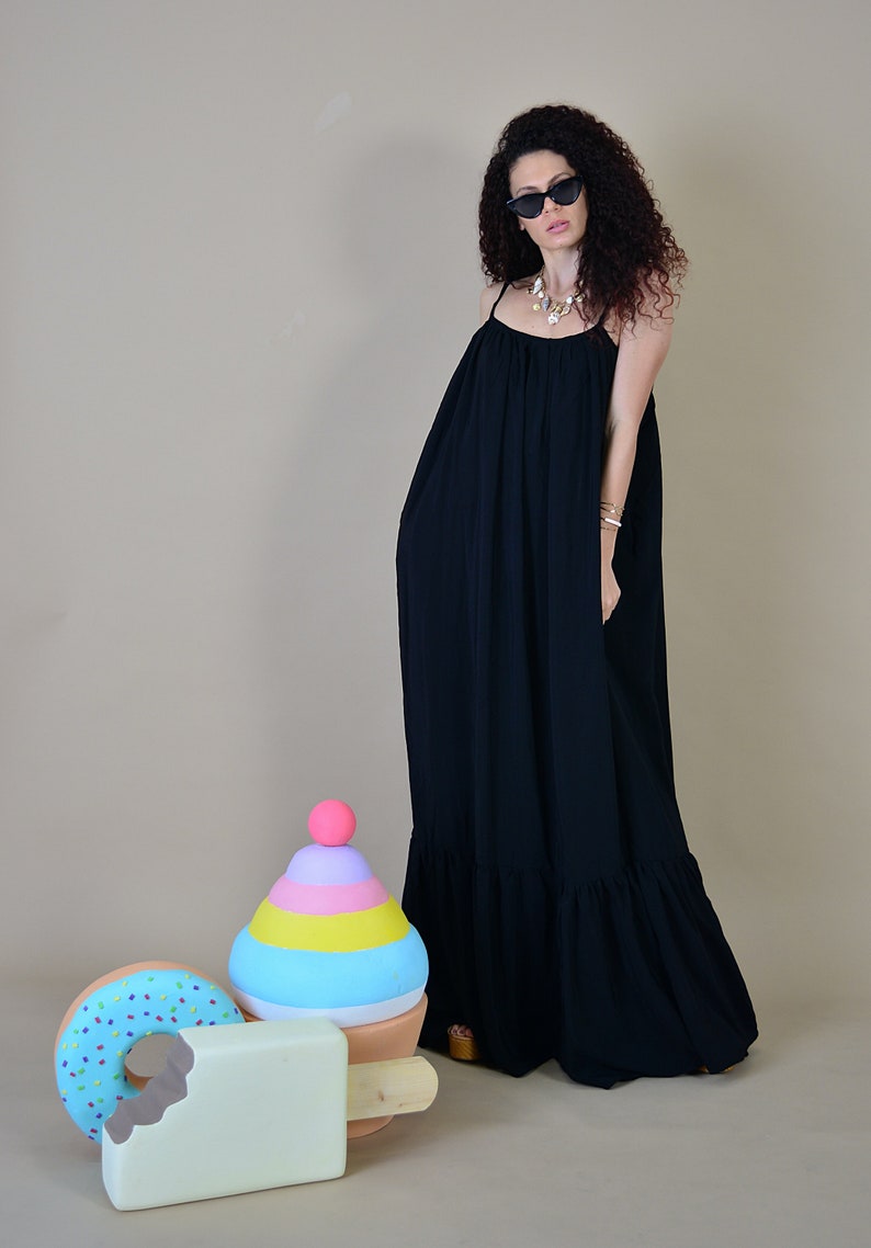 May include: A woman wearing a black maxi dress with spaghetti straps and sunglasses. The dress has a tiered ruffle at the hem. Decorative oversized ice cream, donut, and popsicle props are in the foreground.