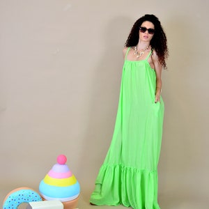 May include: A woman wearing a bright green maxi dress with a ruffled hem and sunglasses. She is standing in front of a light brown background with a colorful toy in the foreground.