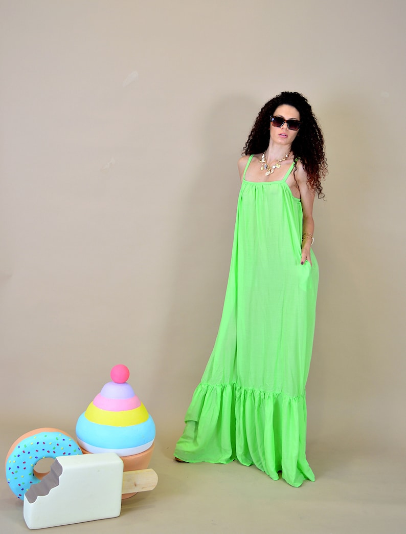 May include: A woman wearing a bright green maxi dress with a ruffled hem and sunglasses. She is standing in front of a light brown background with a colorful toy in the foreground.